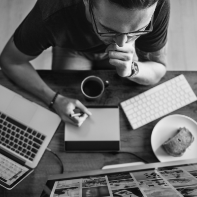 Une personne est assise à un bureau et travaille avec une tablette graphique, entourée d'un ordinateur portable, d'un clavier, d'une tasse à café et d'une assiette avec une tranche de pain, vue d'en haut en noir et blanc.