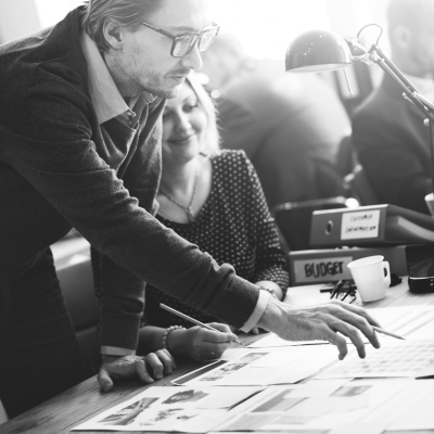 Un homme portant des lunettes est penché sur un bureau, montrant des documents tout en discutant avec une femme. D'autres personnes travaillent à l'arrière-plan. Une étiquette indiquant "BUDGET" est visible sur le bureau. L'image est en noir et blanc.
