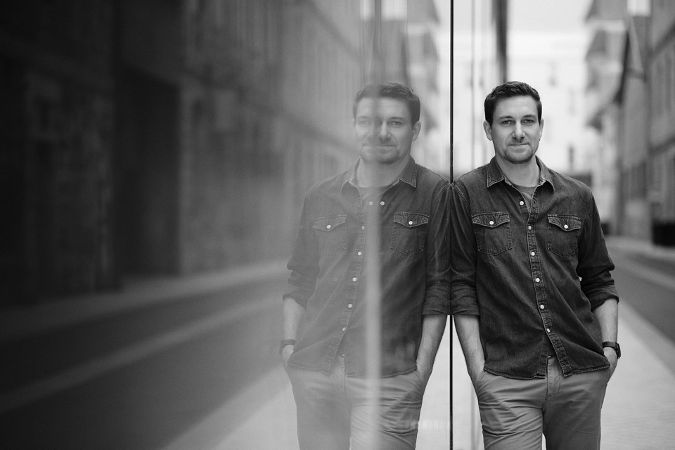 Un homme vêtu d'une chemise en jean s'appuie contre une paroi en verre dans une rue de la ville, son reflet étant visible. L'image est en noir et blanc et la rue semble calme et vide.