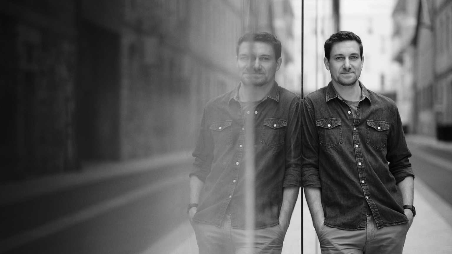 Un homme vêtu d'une chemise en jean se tient de manière décontractée, adossé à une paroi en verre où l'on peut voir son reflet. L'arrière-plan montre une rue urbaine vide en noir et blanc.